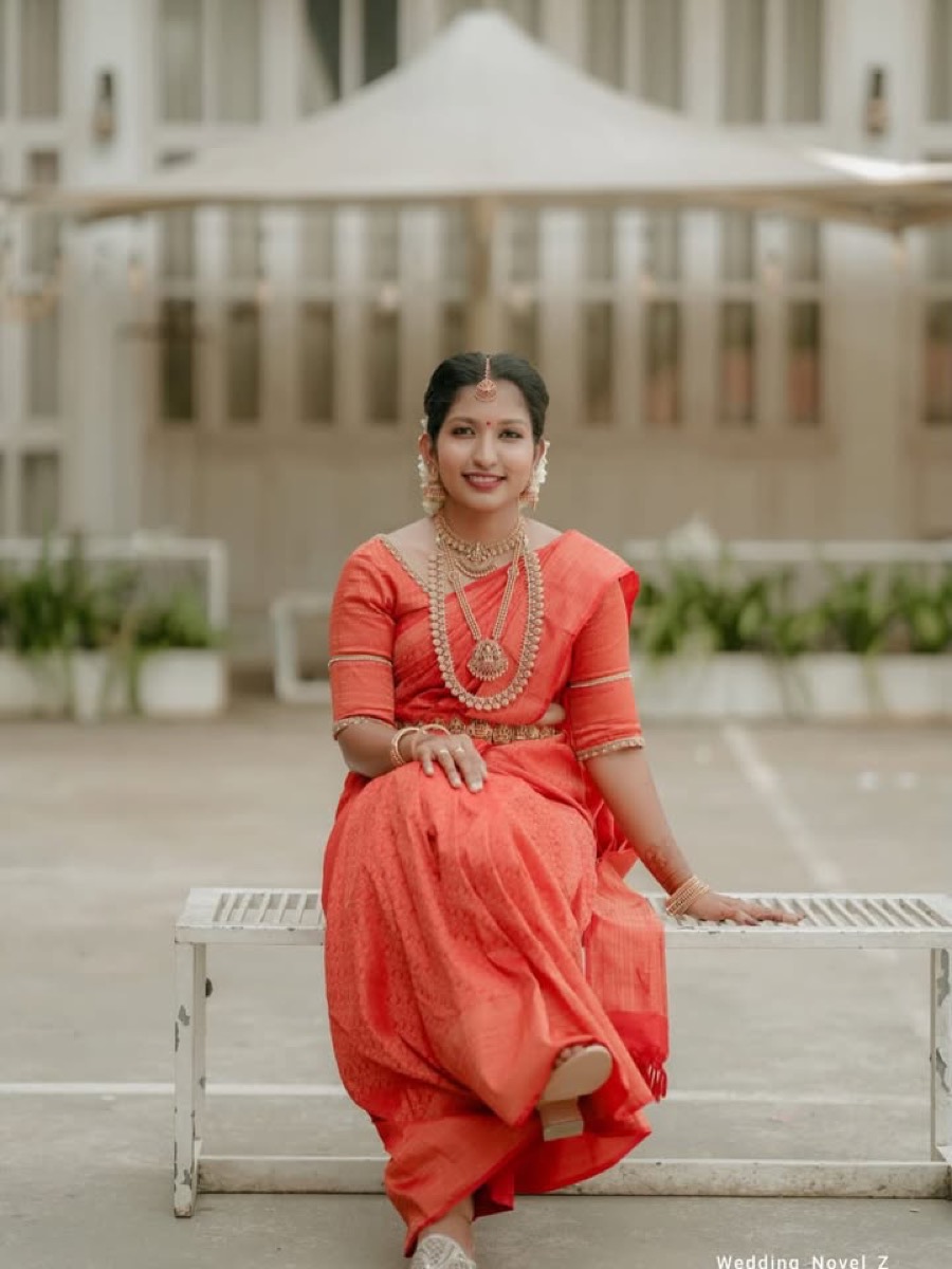 A bride in a coral saree seated on a white bench