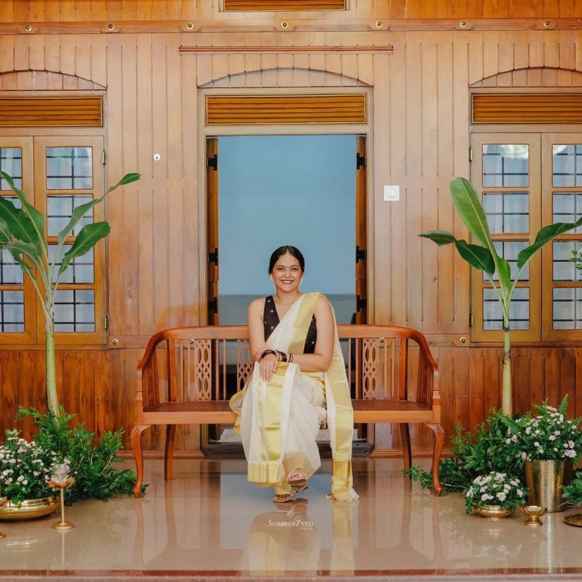 A client in a kasavu set-mundu on a wooden bench, with banana plants and brass lamps