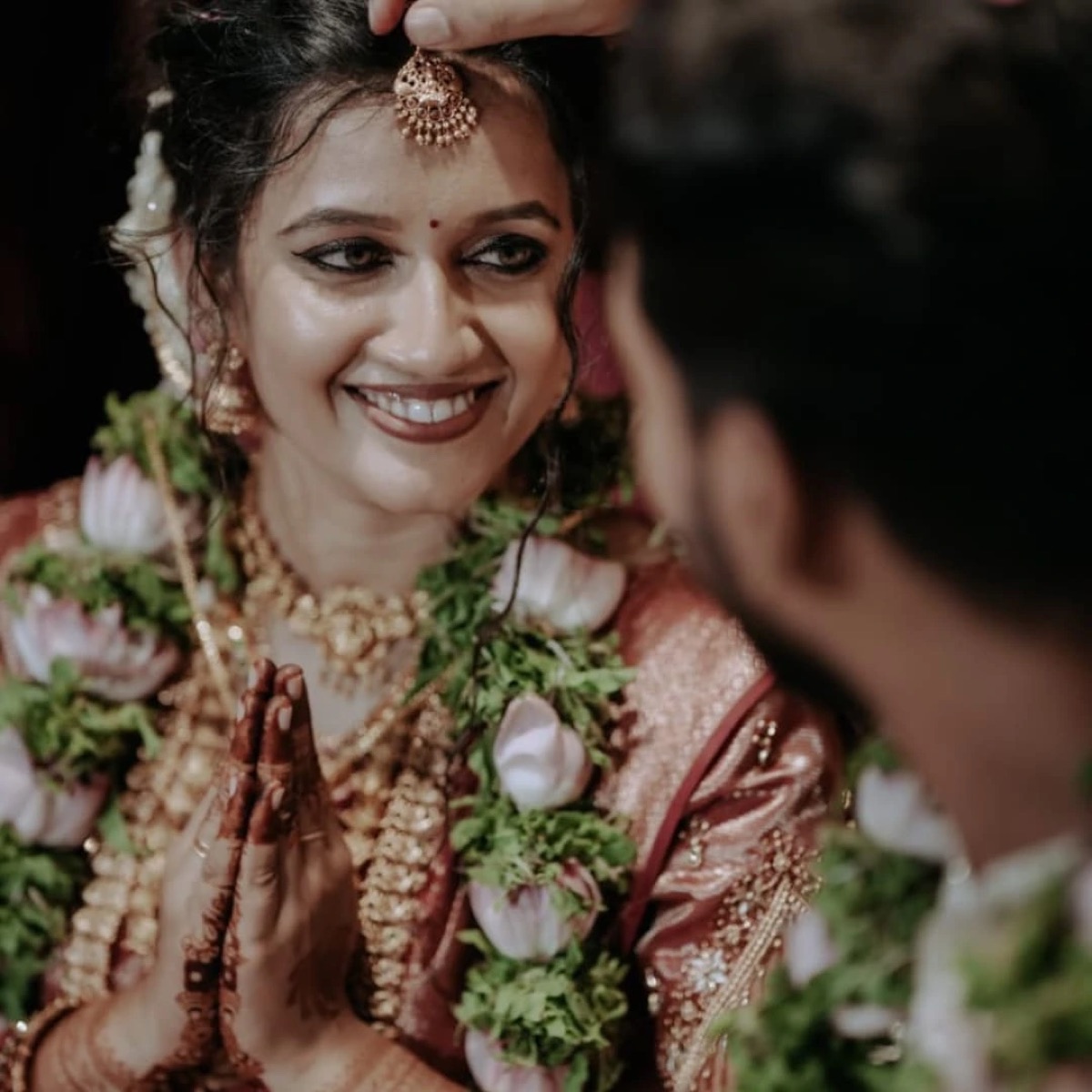 A smiling bride with folded hands at her wedding ceremony, wearing a lotus garland