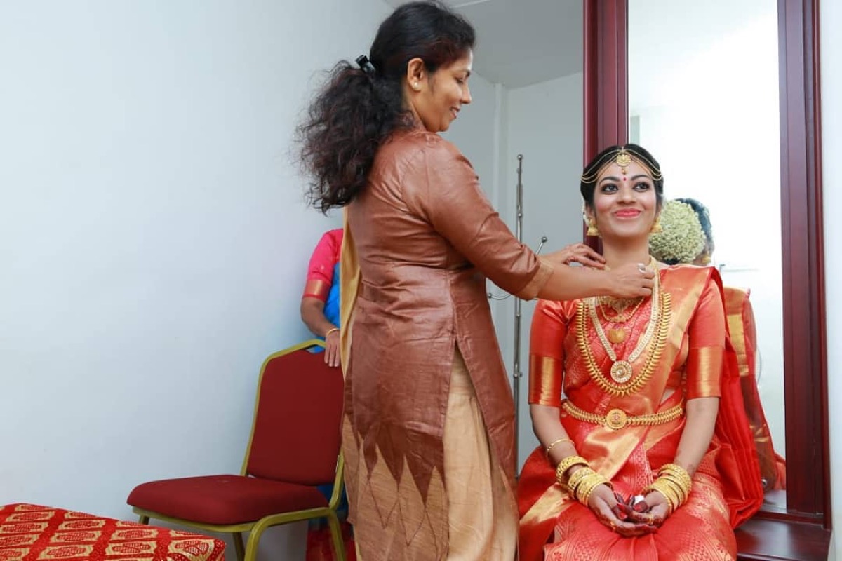 A stylist fixing a seated bride's jewellery in front of a mirror