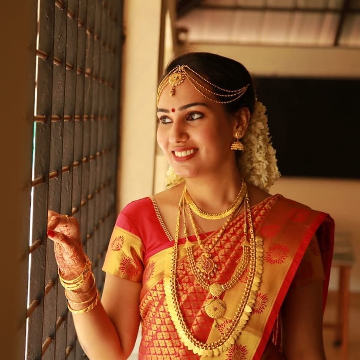 A smiling bride by a window, in red and gold silk with jasmine and layered gold jewellery