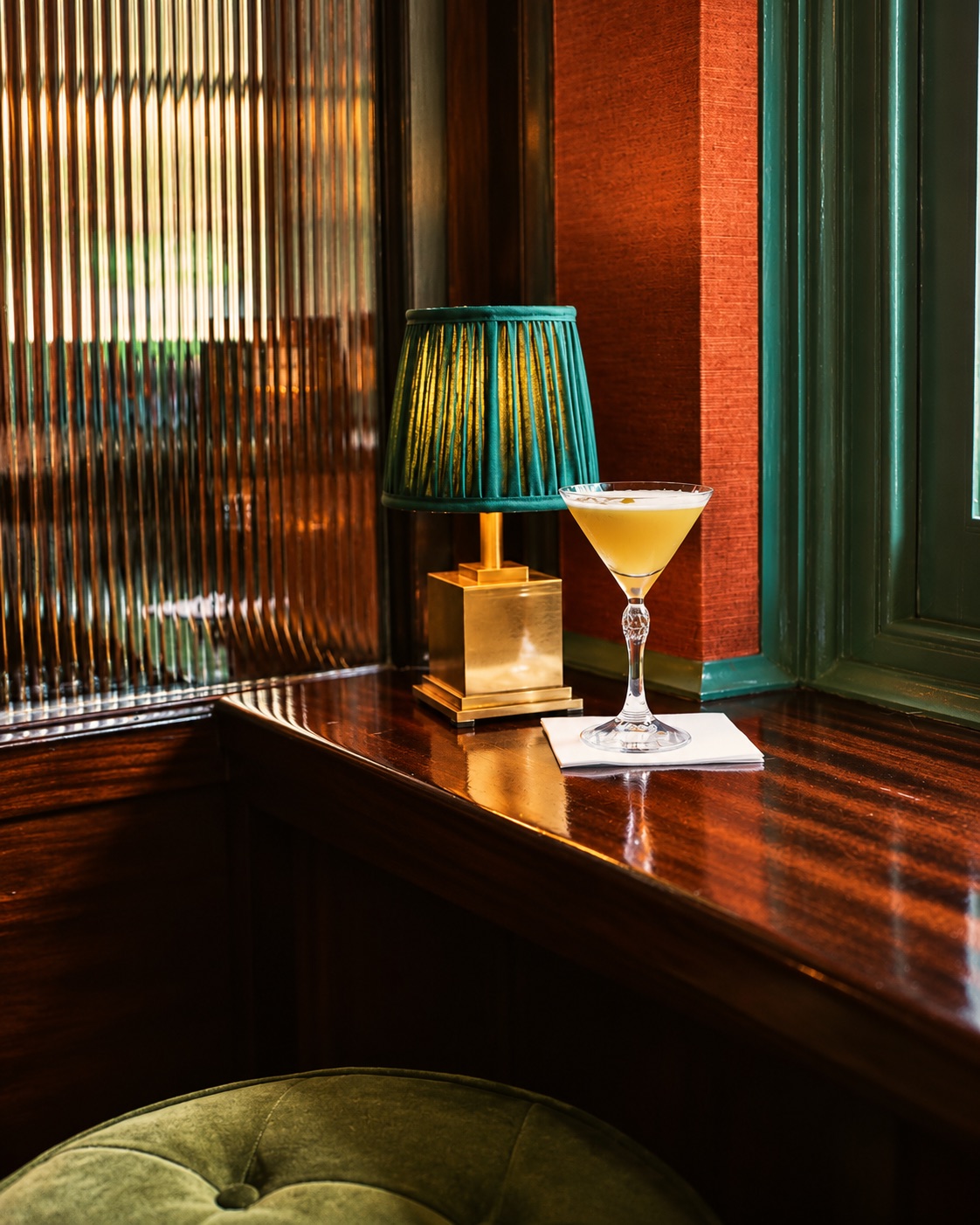 A cocktail beside a green-shaded lamp on a polished bar